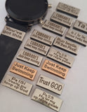 A black smartwatch with a black strap is shown alongside multiple rectangular metal watch tag charms engraved with motivational and religious phrases, arranged on a white surface.