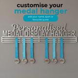 A personalized metal medal display hanger mounted on a gray wall, with six blue ribbon medals hanging below. Large text above reads 'Personalised MEDAL DISPLAY HANGER' with smaller text 'customise your medal hanger' and 'add your name, sport or favourite quote'.