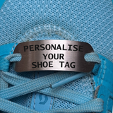 A stainless steel shoe tag with the text 'PERSONALISE YOUR SHOE TAG' engraved on it, placed on a blue shoe with a focus on the tag.
