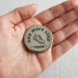 Hand holding a round metal token with 'one more step, repeat' text and a shoe icon.