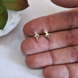 A pair of small stainless steel stud earrings shaped like stylized ballerinas, shown resting on a person's fingers against a light background with a plant leaf in the corner.