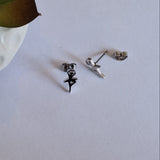 A pair of small stud earrings displayed on a light gray surface, with one earring shown front-facing in black and the other turned sideways showing the silver backing. A white textured ceramic pot with green leaves is partially visible in the top-left corner.