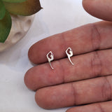 A pair of small silver earrings shaped like stylized figures, displayed on a person's fingers against a light background with a plant pot edge visible.