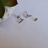 A pair of silver-toned geometric stud earrings with flat, angular shapes, shown disassembled with their posts and backings separated, alongside a small metal component on a light surface next to a white textured planter.
