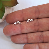 A pair of small silver mountain-shaped stud earrings resting on a person's fingers, with a small plant leaf visible in the upper left corner.