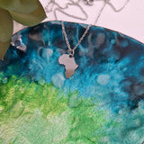 A silver necklace with an Africa-shaped pendant resting on a blue, green, and black watercolor-textured ceramic plate.