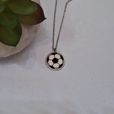 A silver chain necklace with a circular pendant featuring a black and white soccer ball design, displayed on a light surface next to a small green succulent.