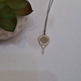 A silver padel racket pendant necklace on a delicate silver chain, displayed on a light gray surface next to a small potted succulent.