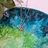 A silver chain necklace with a paw print pendant resting on a glossy, water-droplet-covered ceramic plate with a blue and green gradient.