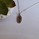 A silver oval pendant necklace with black engraved symbols of a swimmer, bicycle, and running figure, displayed on a light surface next to a small potted succulent.