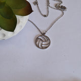 A silver-toned netball pendant necklace on a delicate chain, styled on a white surface next to a green succulent in a white pot.