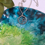 A silver necklace with a circular cutout pendant featuring a netball pattern, displayed on a glossy, watercolor-textured blue and green plate.