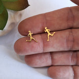 Two small gold stud earrings shaped like running female athletes, displayed on a person's fingers against a white background.