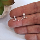 A pair of small silver stud earrings shaped like running female silhouettes, shown resting on a person's fingers for scale.