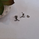 A pair of small silver stud earrings shaped like stylized human figures, shown on a white surface with a decorative leafy object partially visible in the top left corner.