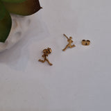 A pair of small gold-tone stud earrings shaped like running figures, shown on a white surface with a green leaf and white textured ceramic object in the background.