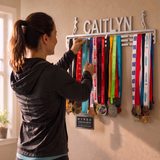 A woman in a black athletic hoodie and shorts hangs a medal on a wall-mounted race ribbon rack personalized with the name CAITLYN. The rack is filled with multiple colorful race ribbons and medals, and a small Hyrox brand plaque is visible.