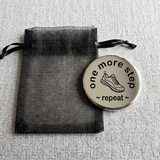 Motivational coin with 'one more step, repeat' text on a black organza drawstring bag.