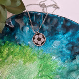 A silver chain necklace with a circular pendant featuring a black and white soccer ball design, displayed on a glossy blue and green textured plate.