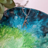 A silver-toned pendant necklace with a person playing netball from a chain, displayed on a glossy blue and green ceramic dish with water droplets.