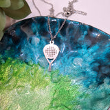 A stainless Steel Padel racket pendant necklace resting on a colorful, textured ceramic plate with blue, green, and black marbled patterns.