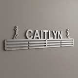A silver metal personalized medal hanger spelling 'CAITLYN' with two small runner silhouettes on either side, mounted on a plain taupe-gray wall.