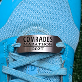 A close-up of a light blue running shoe with a custom metal nameplate engraved with 'COMRADES MARATHON 2027' threaded through the eyelets.