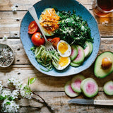 A plate of salad with avocado slices, a fried egg, and tomato, accompanied by a side of toast, a bowl of jam, and a knife, all set on a wooden table with flowers in the foreground.